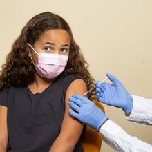 A masked doctor in a white coat and blue gloves administers a vaccine into the arm of a masked child.
