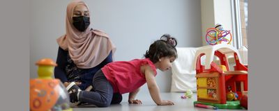 An excited-looking toddler crawls towards toys while her mother watches on.