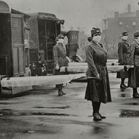 Photograph from 1918 influenza pandemic shows mask-wearing women holding stretchers at backs of ambulances in Saint Louis, Missouri.