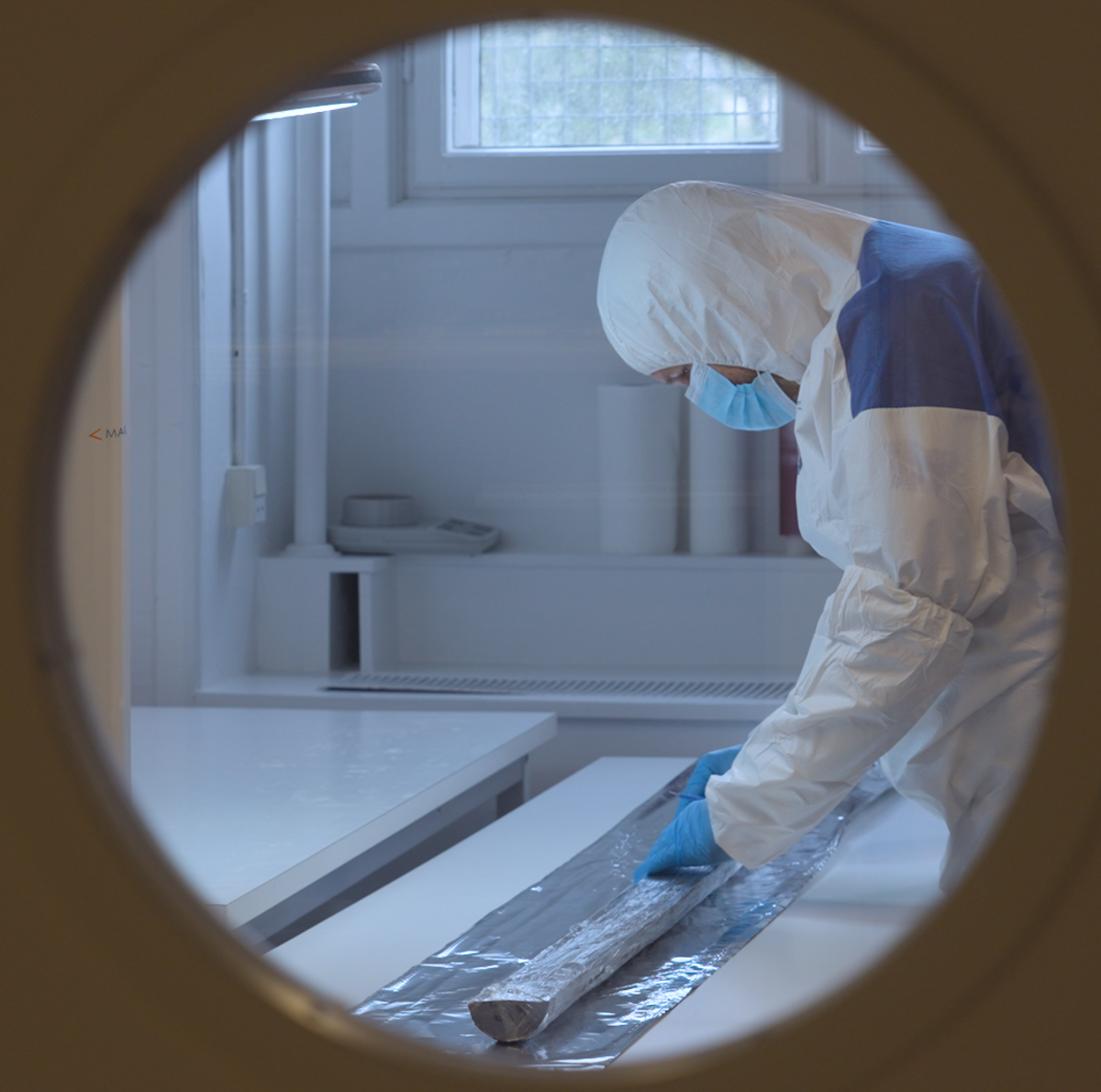 A photo through a door of a researcher in PPE working with a sediment core